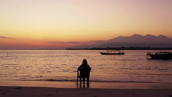 Silhouette of girl sitting on chair over sandy beach, watching colorful sky after sunset with mounta alt