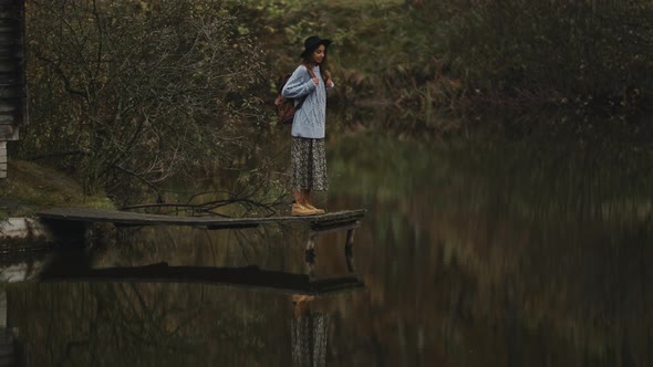 Stylish Inspired Traveler Woman in Hat with Backpack Standing on Wooden Pier Promenade at Lake alt
