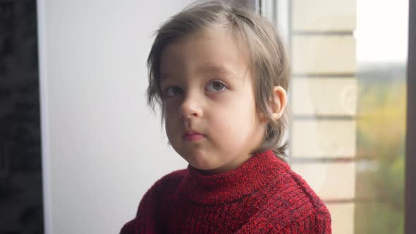 Boy Child in a Red Sweater and a Knitted Hat Sits on a Large White Window alt