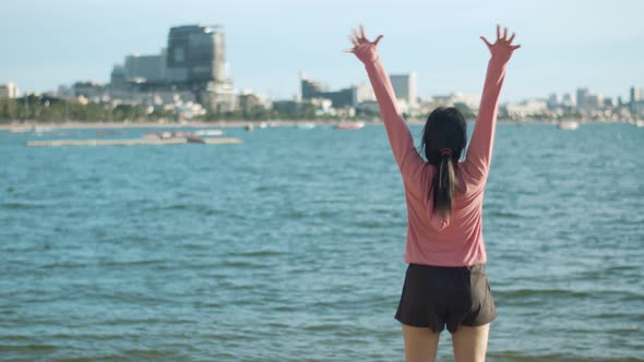 An attractive woman jogging in sportswear raises arms into the air looking at the ocean view. alt