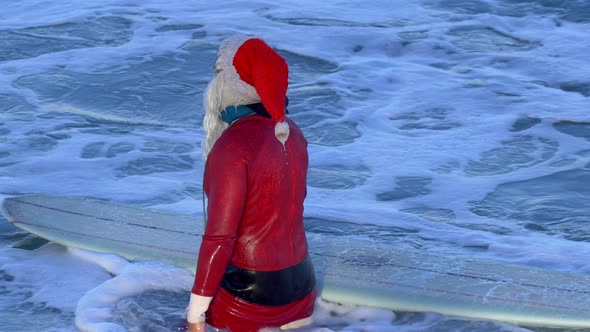 Santa Claus walking with his surfboard to go surfing at the beach. alt