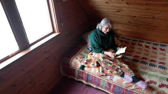 Woman in Old Age Sits on a Bed Under a Blanket with a Book in Her Hand