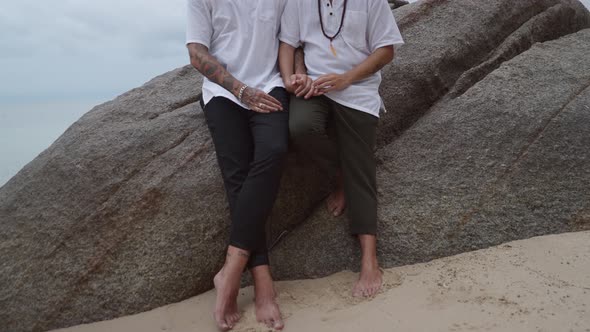 Gay Couple Holding Hands Sitting Close to Each Other on a Big Rock on the Beach No Face Push in Shot alt