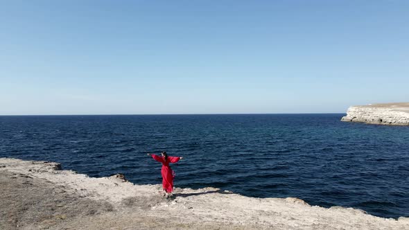 Brunette Woman in a Red Long Dress Stands on the Edge of a Cliff By the Sea alt