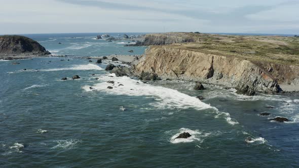 Tracking drone footage of sea cliffs at the Pacific ocean, on a sunny day. Waves crash against the r alt
