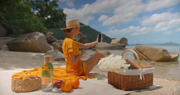 Nice Woman in Yellow Dress and Hat Having Video Call By Her Phone on Sunny Day Sitting on the Beach alt