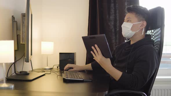 A Young Asian Man in a Face Mask Works on a Desktop Computer and a Tablet at Home alt