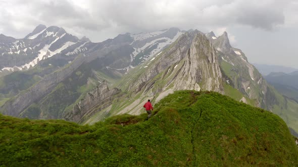A man running on top of a mountain. He is wearing a red jacket. To his left and right it goes down a alt