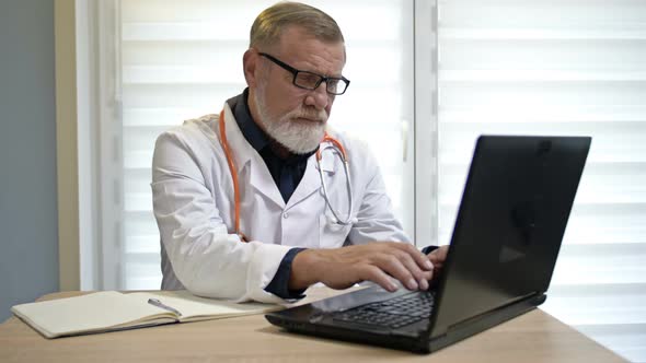 Elderly Doctor Works at a Computer in His Office. Modern Technologies and Many Years of Experience. alt