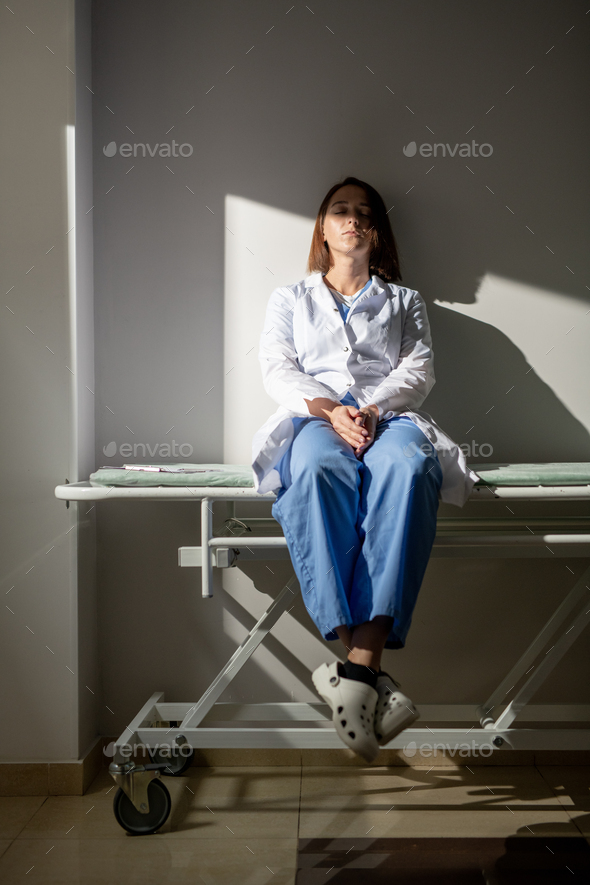 Young exhausted nurse of covid hospital in whitecoat sitting on medical cart