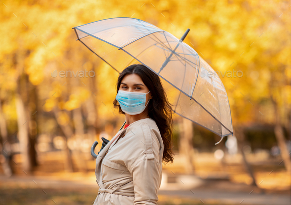Pretty young woman in surgical mask on walk under rain at park on fall day
