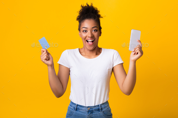 Excited black lady holding credit card and cellphone at studio