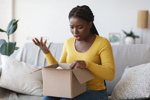 Wrong Parcel. Shocked frustrated black woman unpacking box, suffering from shipping mistake
