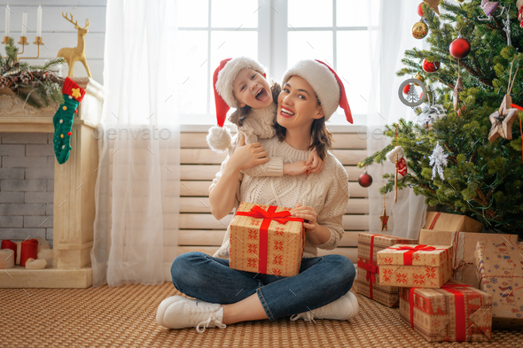 Merry Christmas and Happy Holidays! Cheerful mom and her cute daughter girl exchanging gifts. Parent and little child having fun near tree indoors. Loving family with presents in room.