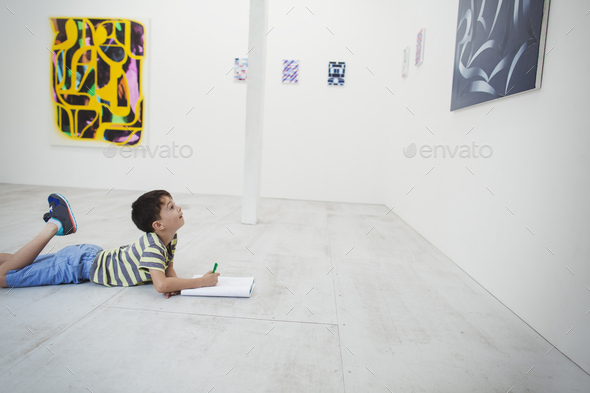 Boy lying on floor in art gallery with pen and paper, looking at modern painting.