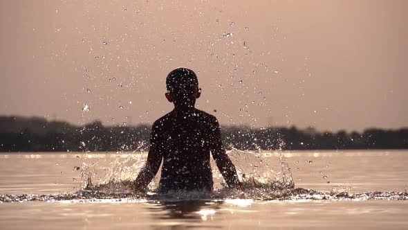 Silhouette of Happy Boy at Sunset Creating Splashes of Water with His Hands. Slow Motion alt