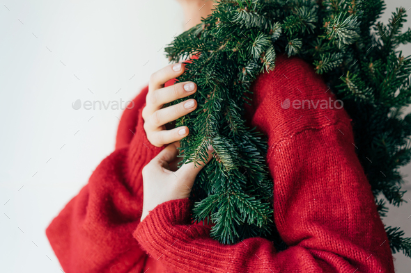 A natural spruce wreath hangs on the shoulder of a woman in a red sweater.