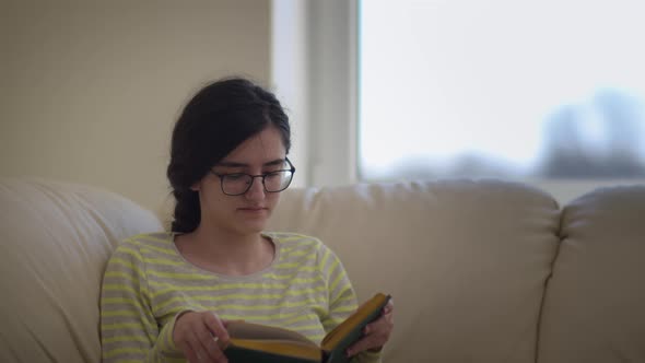 Pretty Girl with Glasses Sitting on the Sofa Reading a Book and Smiling