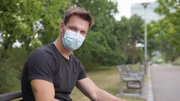 A Young Man Points at the Camera and Nods As He Sits on a Bench in a Park - Closeup From the Side alt