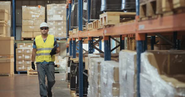 Logistics worker wearing a helmet working in a large warehouse checking inventory alt