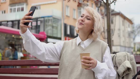 Young Blonde Business Woman in a Beige Vest and White Blouse Sits on a Bench in the Middle of the alt