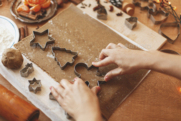 Hands cutting gingerbread dough with man, star and tree metal cutters on rustic table with spices, festive decorations, lights. Person making Christmas gingerbread cookies, holiday advent