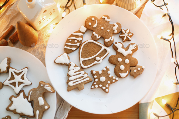 Decorated christmas tree, star and reindeer cookies with sugar frosting, flat lay. Gingerbread cookies with icing on white plate on wooden table with festive lights. Christmas holiday tradition