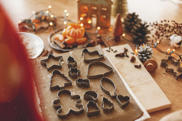 Making gingerbread cookies, Christmas holiday tradition. Raw gingerbread dough with metal cutters, spices, oranges, festive decorations and lights on rustic table.