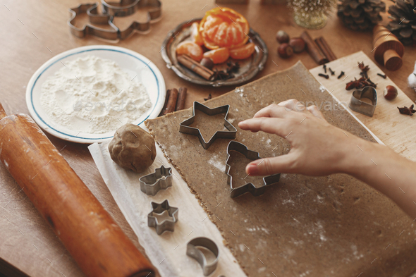 Hands cutting gingerbread dough with festive star and tree metal cutters on rustic table with spices, festive decorations, lights. Person making Christmas gingerbread cookies, holiday advent