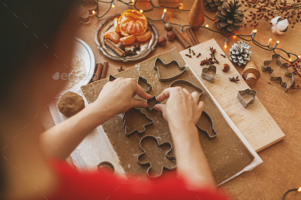 Person making Christmas gingerbread cookies, holiday advent. Hands cutting gingerbread dough with festive metal cutters on rustic table with spices, oranges, festive decorations, lights