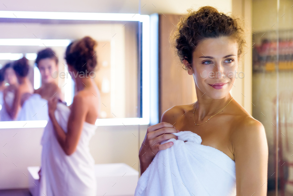 Smiling woman reflected multiple times in a mirror as she poses in a clean white towel in the bathroom preparing for a shower