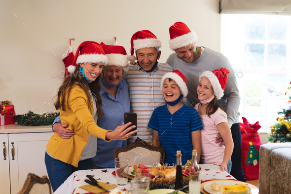 Multi generation caucasian family standing at table for dinner together, wearing santa hats and face masks, taking a selfie and smiling. quality family time christmas celebration.