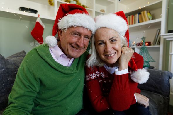 Portrait of senior couple in santa hat smiling while sitting on couch at home. social distancing during covid 19 coronavirus quarantine lockdown.