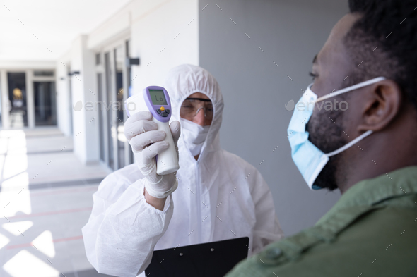 African American businessman is shown his temperature by worker in protective suit before being allowed in to office. Hygiene and social distancing in workplace during Coronavirus Covid 19 pandemic.