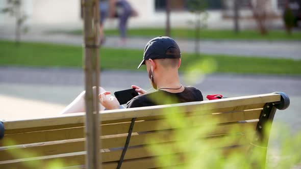 The Young Man Sits in the Park and Chats on the Smartphone. Happy Man Use Smartphone Sitting on alt