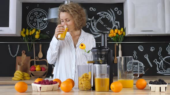 Young Blonde Woman In Pajamas Drinking Fresh Orange Juice In Kitchen alt