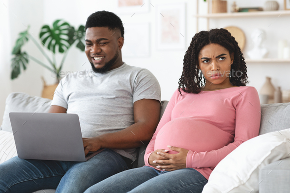 Angry pregnant african woman sitting by her husband using laptop