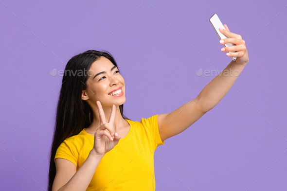 Selfie Fun. Cheerful Asian Woman Taking Self-Portrait On Smartphone, Showing Peace Gesture