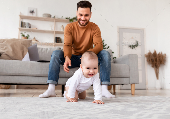 Happy Daddy Looking At Baby Crawling On Floor At Home