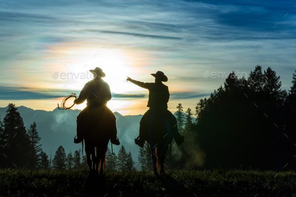 Two cowboys riding across grassland with mountains in background, early morning - Stock Photo - Images
