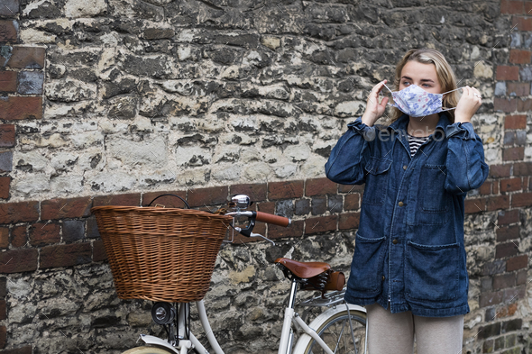 Young blond woman standing next to bicycle with basket, putting on face mask.