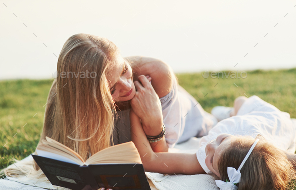 Mom and daughter reading a book in a sunny day laying on the grass