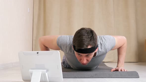 Young Man with a Bandage on His Head Looks Into a Tablet and Clumsily Does Pushups on the Floor alt