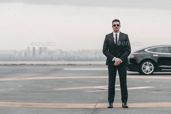 serious bodyguard standing with sunglasses and security earpiece on helipad and looking at camera - Stock Photo - Images