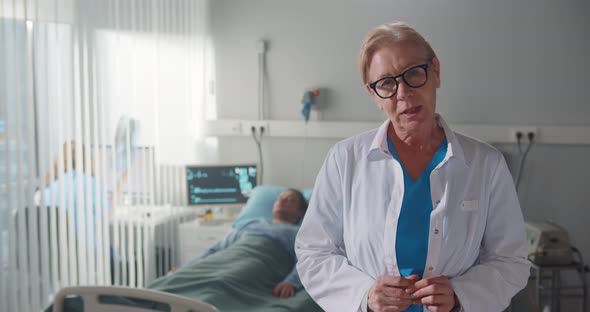 Senior Female Doctor in White Coat Standing in Hospital Ward and Reporting on Patient Treatment alt