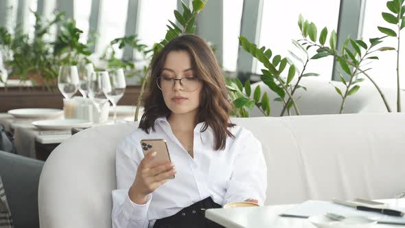 Beautiful Businesswoman Holding Cup of Coffee and Looking at Screen of Mobile Phone Chatting