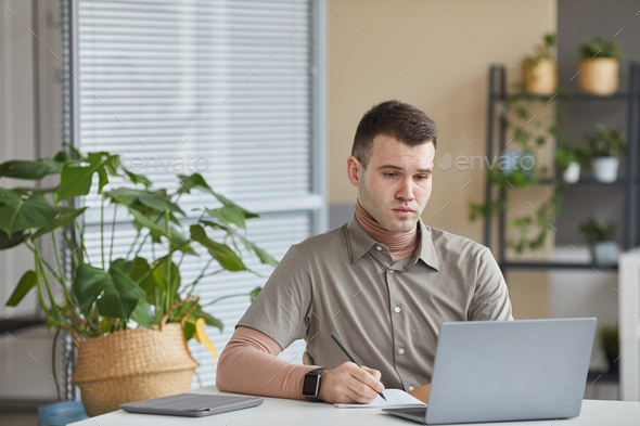 Young office worker sitting at his workplace looking at computer monitor and making notes in note pad