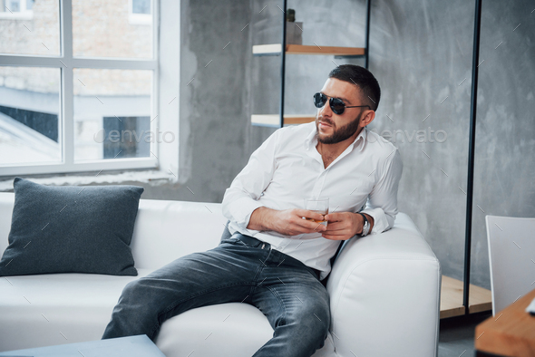 Thoughts about future. Young short-haired man in sunglasses sitting on couch in the office