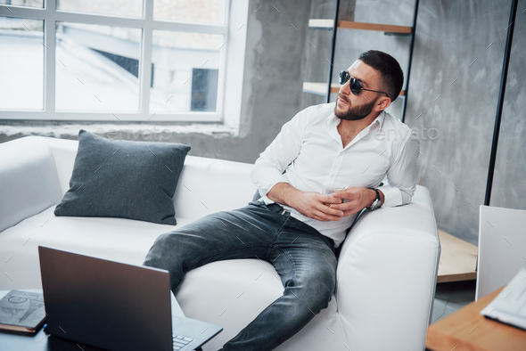 Laptop in the front. Young short-haired man in sunglasses sitting on couch in the office