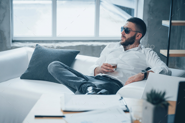 Elegant posing. Young short-haired man in sunglasses sitting on couch in the office